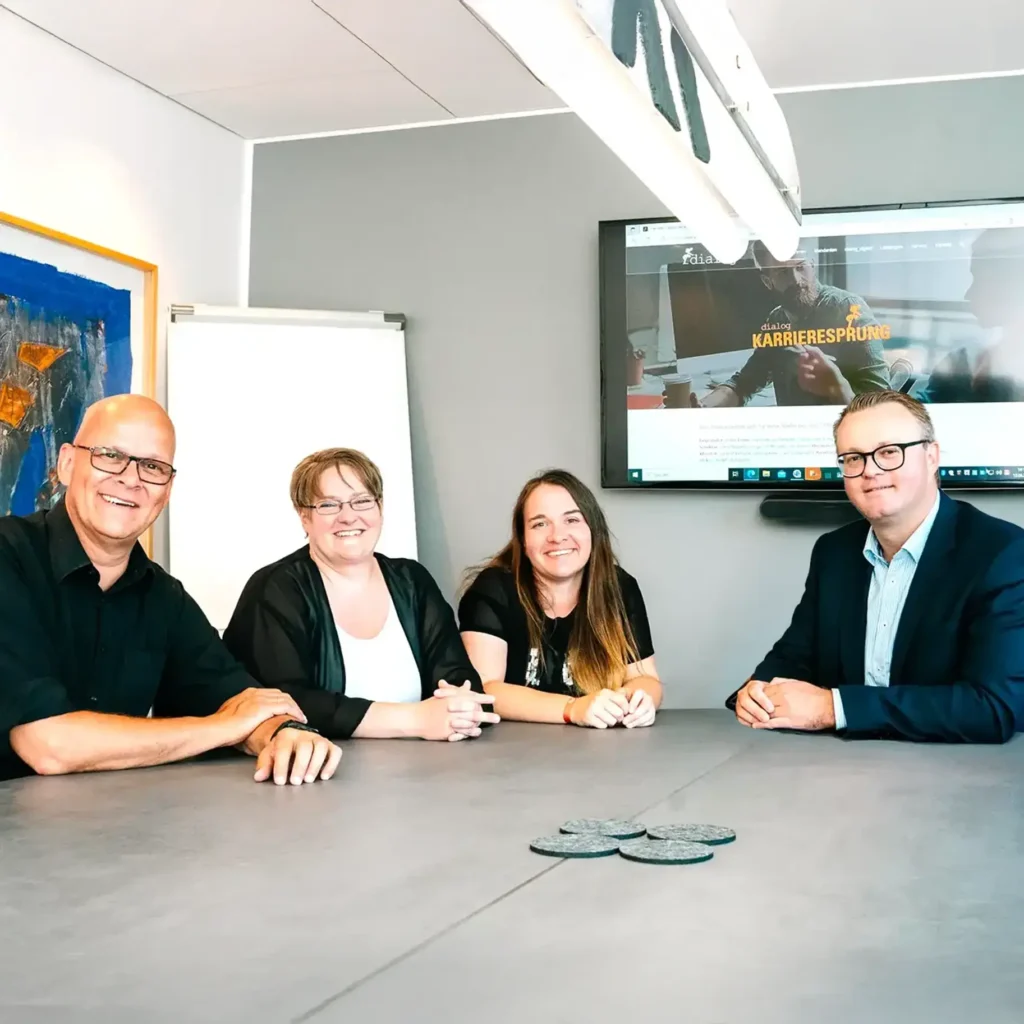 Vier Personen, drei Männer und eine Frau, sitzen an einem Konferenztisch und lächeln in die Kamera. In der modernen Büroumgebung sind im Hintergrund ein Flipchart und ein Bildschirm mit der Aufschrift Lohnabrechnung für Steuerberater und Unternehmen zu sehen.
